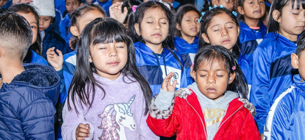 Handsraised_WEB_2025_01_CHIJA3_InicioDeClases-12 Young children, many in Impact School uniforms praying with eyes closed and hands raised