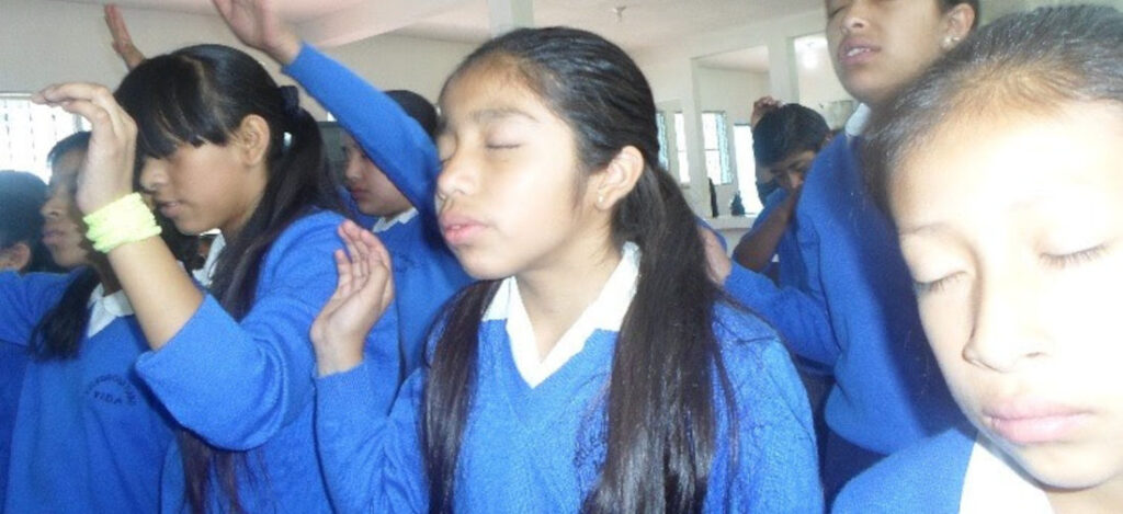 A Guatemalan girl wearing a blue school sweater with hands raised and eyes closed worshiping with classmates