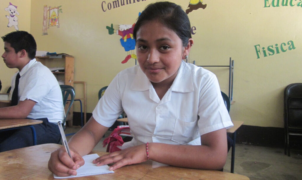 Middle school Guatemalan girl in white button up blouse sitting at a desk and writing on a piece of paper